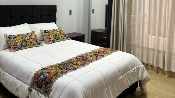 A bedroom with a bed, a nightstand, a mirror, and a window with curtains. - Comfortable/Cozy SKY HOUSE CUSCO (CUSCO)