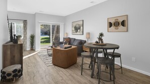 Un salon moderne avec un canapé, une table basse et un coin repas équipé de tabourets. Une grande fenêtre offre une vue sur un jardin.