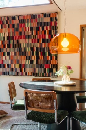 A dining area with a round table, chairs, a hanging pendant light, and a colorful abstract wall hanging. - Private Lakefront Sanctuary | Cedar Hot Tub + 6 Acres, No Neighbors (Nevada City)