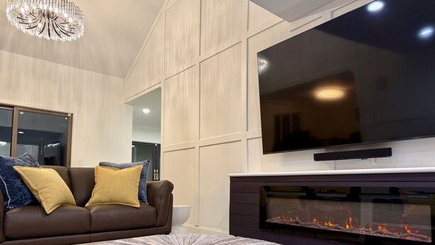 Una sala de estar moderna con un televisor de pantalla plana grande montado en la pared, una chimenea con frente de vidrio, un sofá de cuero marrón con almohadones decorativos y una mesa de centro redonda de madera.
