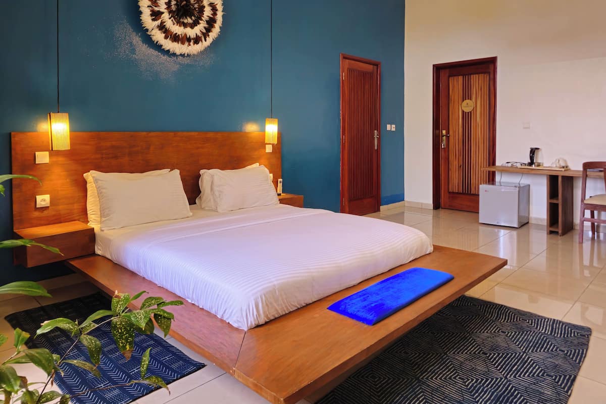 A hotel room with a large bed, a desk with a chair, a small refrigerator, and a wall-mounted lamp.
