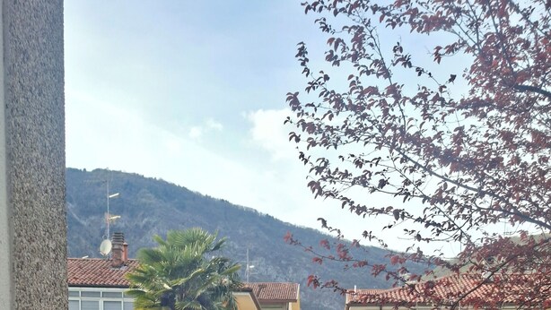 A balcony with two wooden chairs, a small table with a plant, and a view of buildings and hills in the background. - S.I.M residence (MANIAGO)