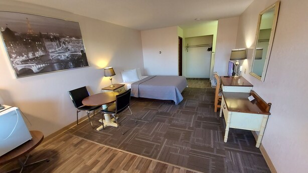 A hotel room with a bed, desk, chair, and a desk lamp. There is a mirror on the wall and a picture of a cityscape. - HOO - DOO MOTEL (Drumheller)
