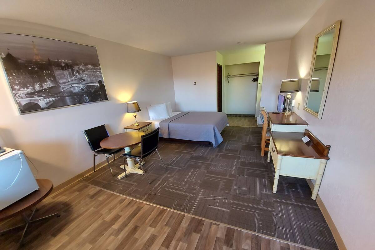 A hotel room with a bed, desk, chair, and a desk lamp. There is a mirror on the wall and a picture of a cityscape.