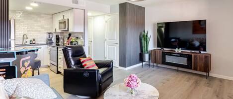 A modern living room with a black leather sofa, a round coffee table with a vase of flowers, a flat-screen TV, and a kitchen in the background.