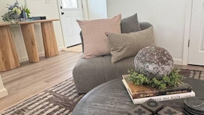 A living room with a grey sofa, a round coffee table with a book and decorative blocks, a patterned rug, and a wooden side table with plants. - Spacious 3BR Home - Fast Wifi - Parking - BBQ - Patio - Garden (Williston)