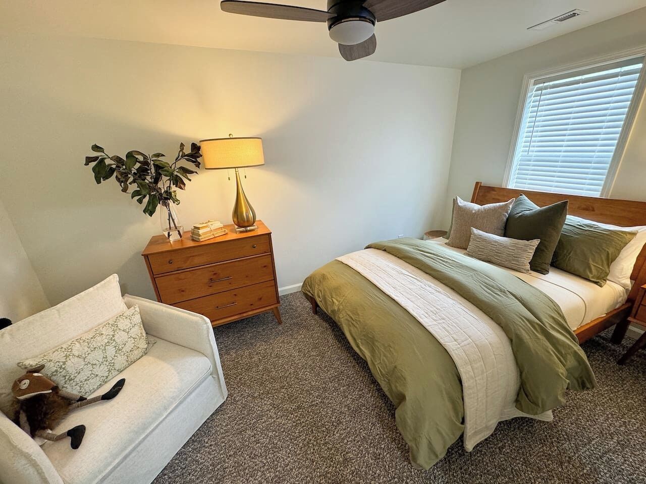 A bedroom with a bed, a dresser, a lamp, a plant, and a cat on a white sofa.