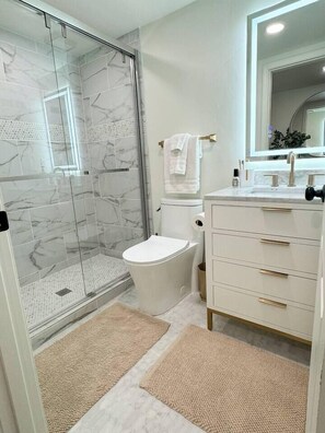 A modern bathroom with a glass shower enclosure, white marble tiles, a toilet, a sink with gold handles, and a mirror above the sink. - Spacious 3BR Home - Fast Wifi - Parking - BBQ - Patio - Garden (Williston)