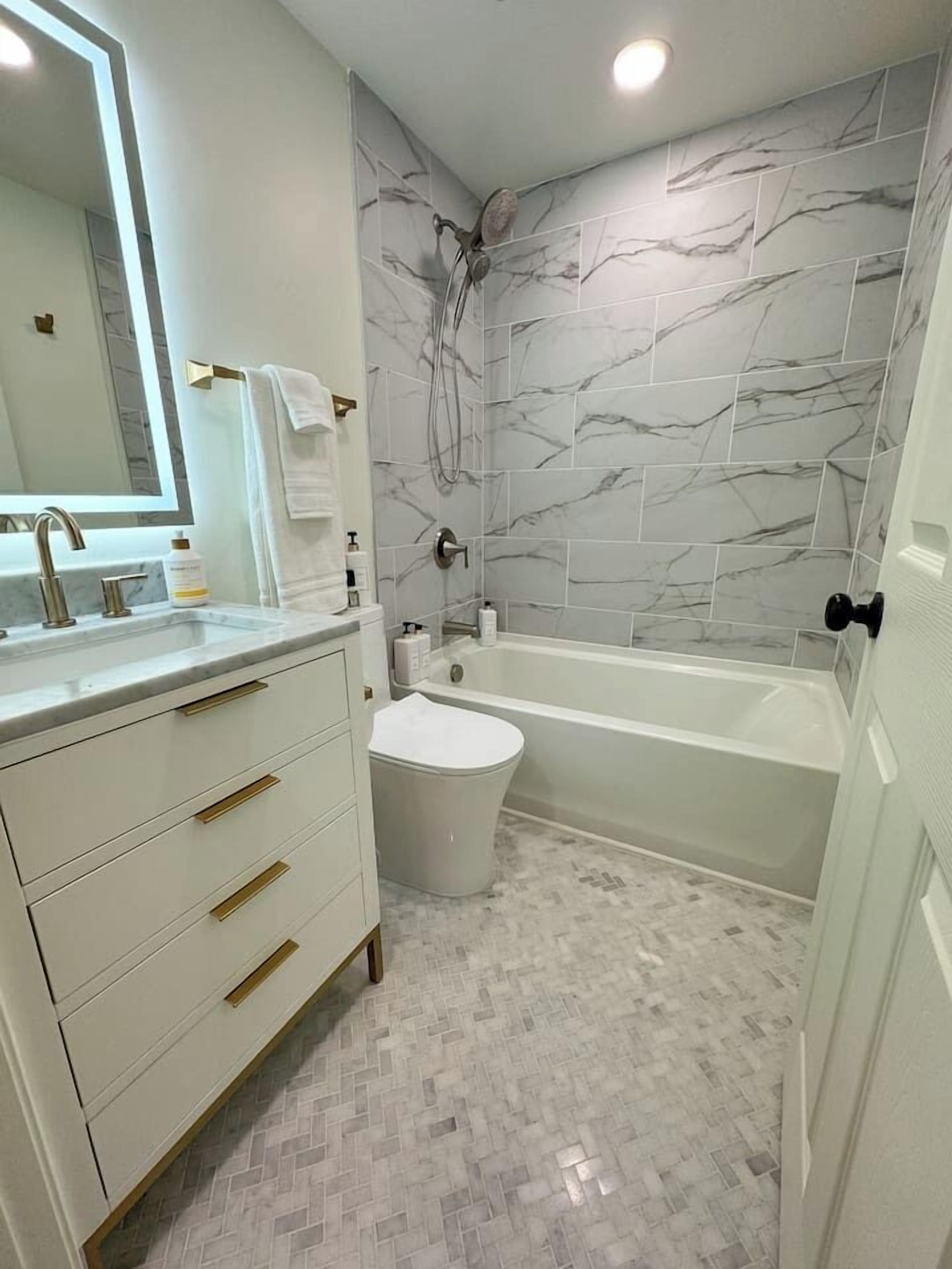 A modern bathroom with a marble shower wall, a freestanding bathtub, and a vanity with gold handles.