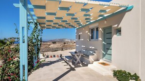 A covered outdoor seating area with a table and chairs, a white building with a blue door, and a view of the landscape.