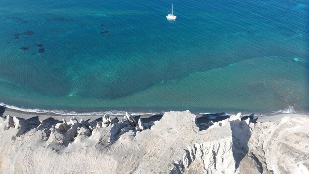 An aerial view of a beach with clear turquoise waters, a small boat anchored near the shore, and rocky cliffs.