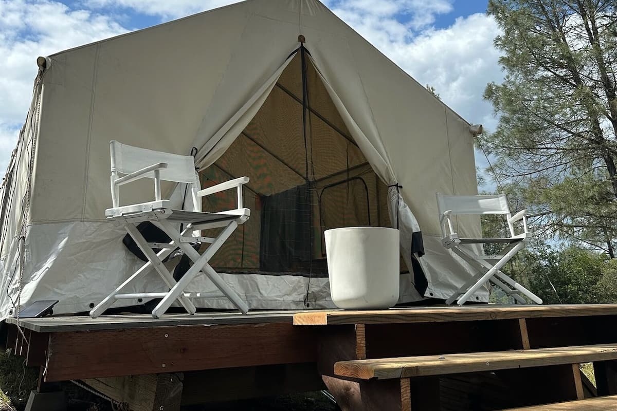 A tent with a flat roof, two folding chairs, a white bucket, and a wooden bench.