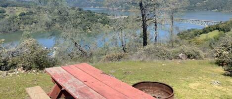Una mesa de picnic con un pozo de fuego en un entorno natural, con un lago y colinas al fondo.
