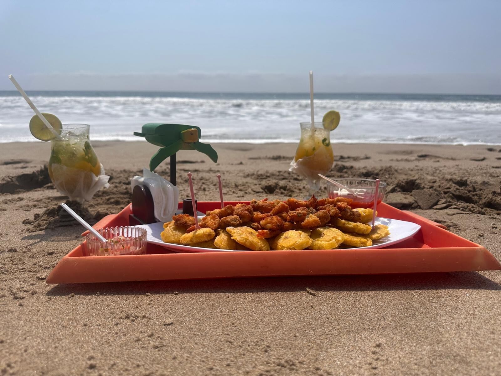 Een schaal gebakken plantanen en twee drankjes op een strand, met de oceaan op de achtergrond.