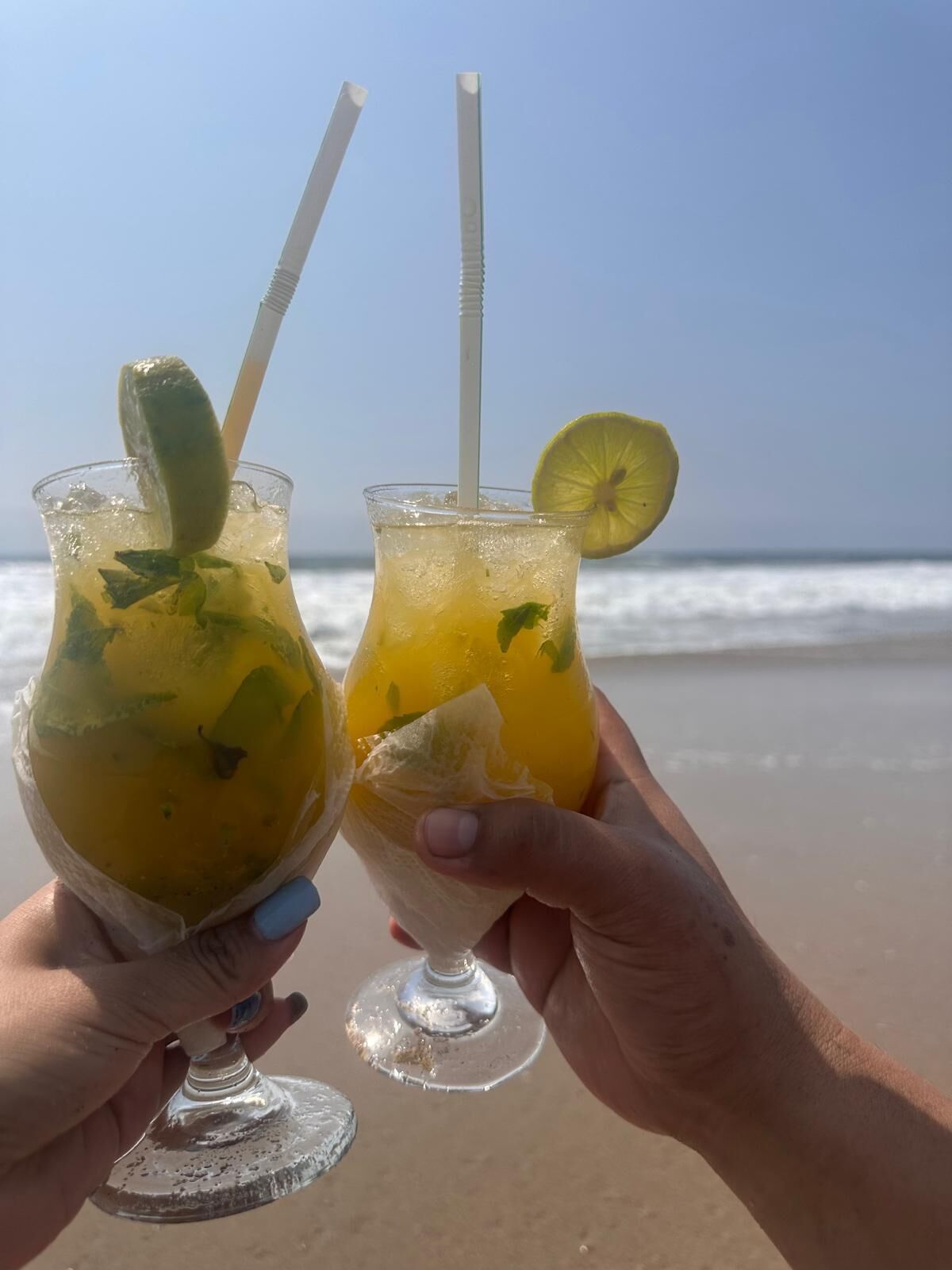 Two drinks with straws and lime slices on a beach.