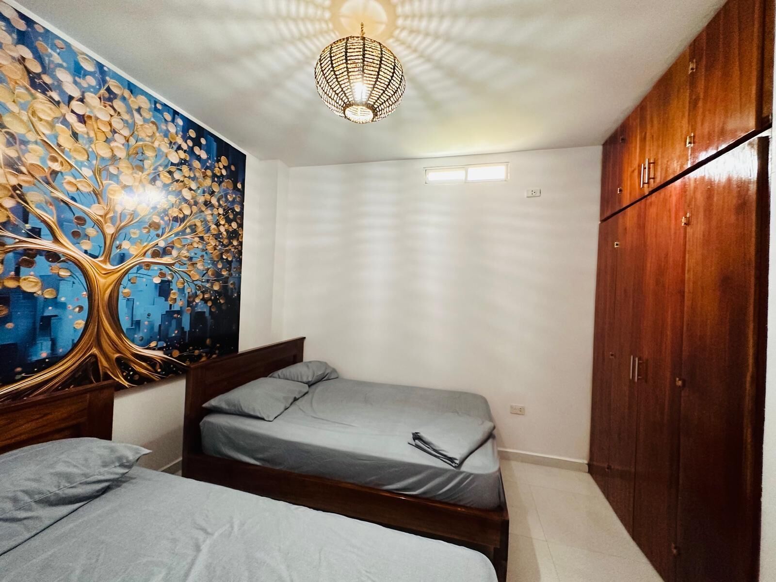 A bedroom with two single beds, a wooden wardrobe, a ceiling light fixture, and a wall mural.