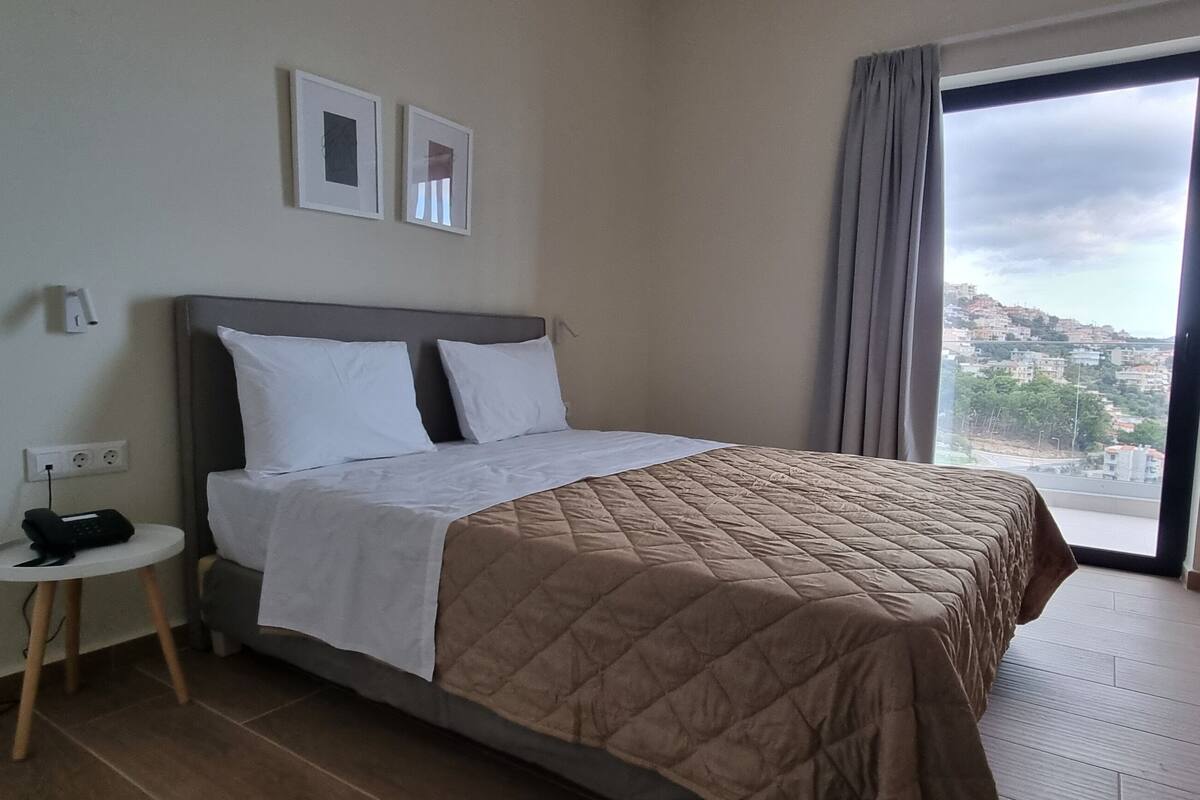 Ein Hotelzimmer mit einem Bett, einem kleinen Tisch mit Telefon, einem Sessel und einem Fenster mit Blick auf Gebäude.