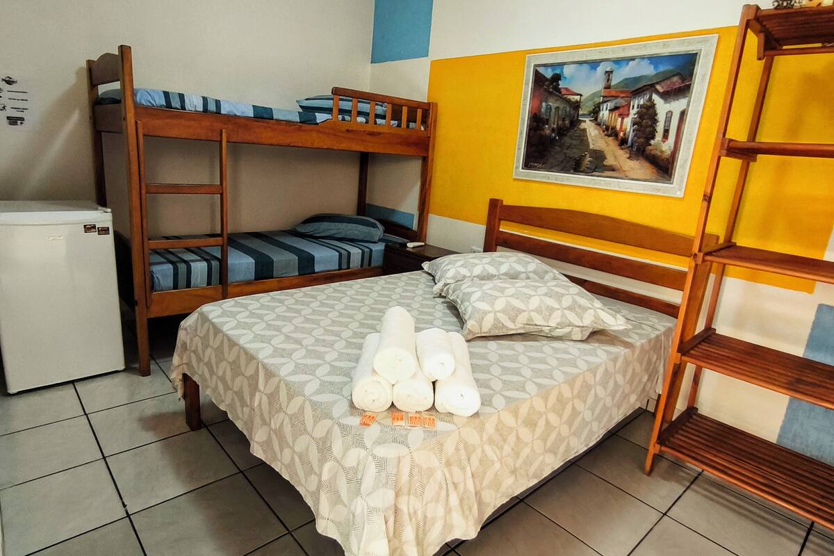 A room with a bunk bed, a small refrigerator, a bed with a patterned bedspread and pillows, and a painting on the wall.