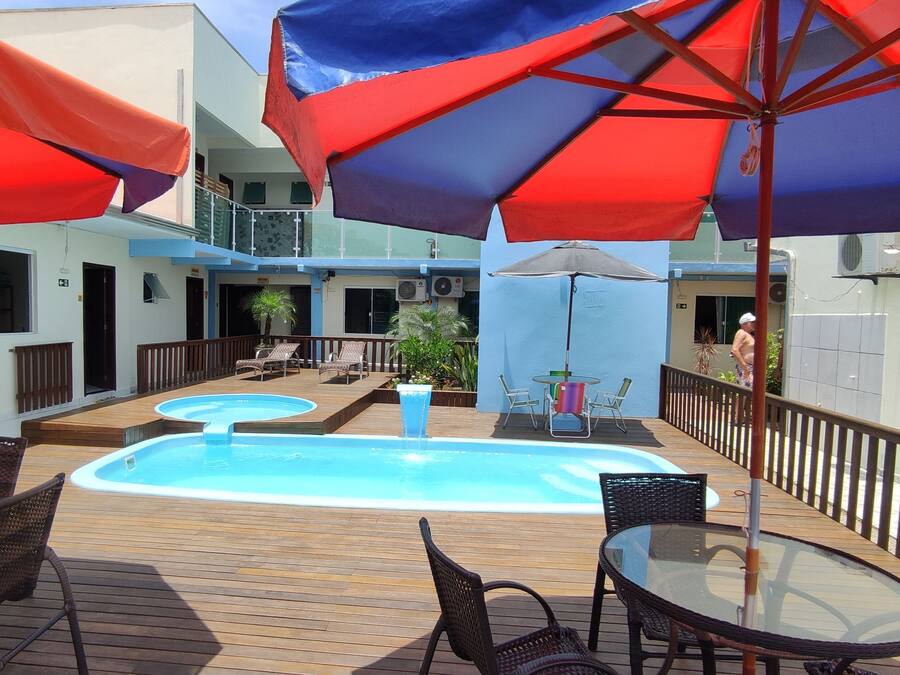 A pool area with multiple lounge chairs and umbrellas, surrounded by a wooden deck and a building with balconies.