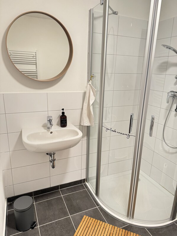 Un baño con lavamanos, espejo, ducha y un cubo de basura.