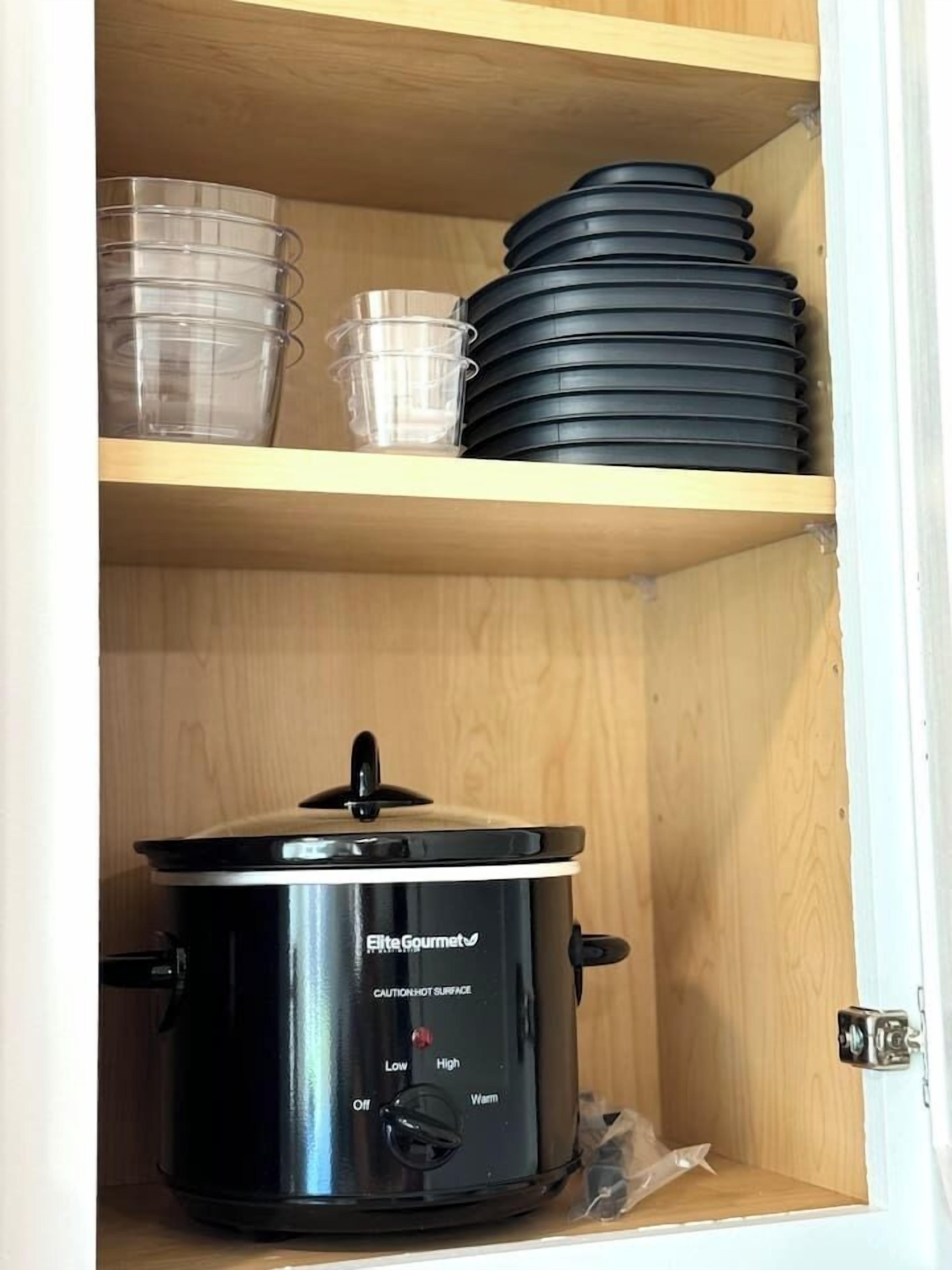 A kitchen with a black Elite Gourmet slow cooker on the counter and a wooden shelf above holding plastic containers and black lids.