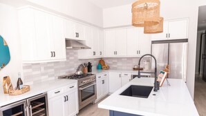 A modern kitchen with white cabinets, a stainless steel range, a built-in oven, a large island with a sink, and a hanging light fixture. - Luxury Beachside Villa - Steps to the sand and beach (Huntington Beach)