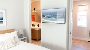 A bedroom with a bed, a nightstand, a lamp, a bathroom with a sink and mirror, and a large ocean view painting. - Luxury Beachside Villa - Steps to the sand and beach (Huntington Beach)