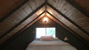 A bedroom with a sloped ceiling, a bed with white bedding, a window with a view of greenery, and a wall-mounted lamp.