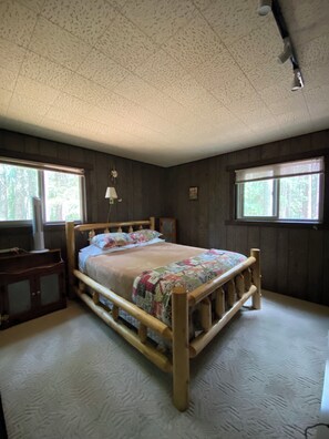 A bedroom with a wooden bed, two windows, a nightstand, and a lamp.