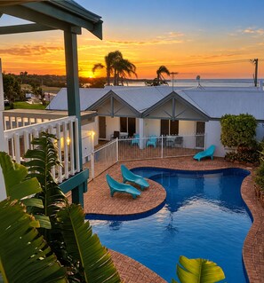 A swimming pool with lounge chairs, a covered patio, and a view of the sunset over the ocean.