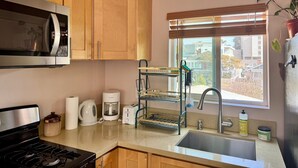 Une cuisine avec des meubles en bois, un four à micro-ondes en acier inoxydable, une machine à café, un grille-pain, un évier et une fenêtre avec des stores.