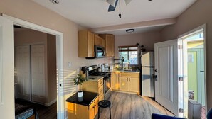 Une cuisine compacte avec des meubles en bois, un four à micro-ondes et un réfrigérateur. On y trouve un ventilateur de plafond, un petit îlot avec une plante et un coin repas avec une chaise.