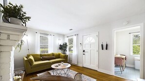Een ruime woonkamer met een groene bank, een ronde houten salontafel en een eethoek met een roze stoel.