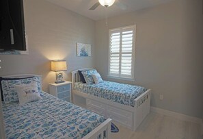 A bedroom with two single beds, a nightstand with a lamp, a ceiling fan, and a window with blinds.