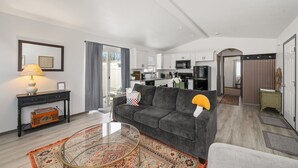 Een moderne woonkamer met een grijze bank, een glazen salontafel en een kamerkleed met een patroon. Achtergrond is een keuken zichtbaar met witte kasten en een magnetron.