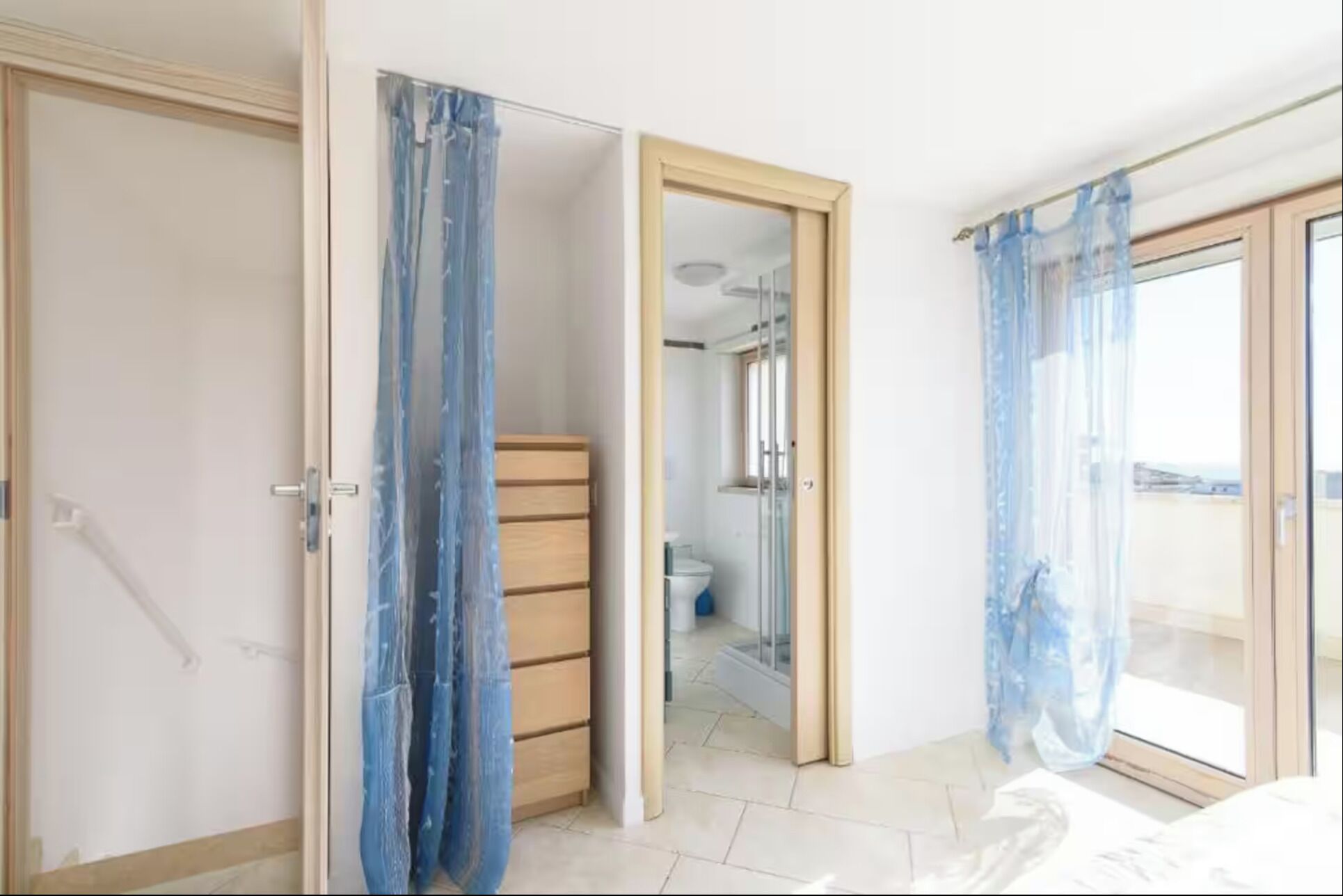 A room with a wooden wardrobe, a bathroom with a toilet and shower, and a balcony with a view.