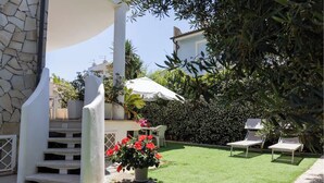 Un espace patio agrémenté d’un mur en pierre, d’une colonne blanche, d’un parasol blanc, d’une petite table décorée de fleurs rouges et de deux fauteuils de jardin.