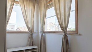 Une pièce dotée de deux fenêtres, chacune ornée de rideaux beiges maintenus par des liens, laissant entrevoir une vue sur les bâtiments à l’extérieur. Un banc blanc est placé contre le mur, sous les fenêtres.