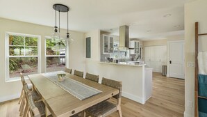 A modern kitchen with white cabinets, a central island, and a dining area with a wooden table and chairs. - NEW Secluded Hillside Retreat on 10 acres with Stunning Views (Port Angeles)