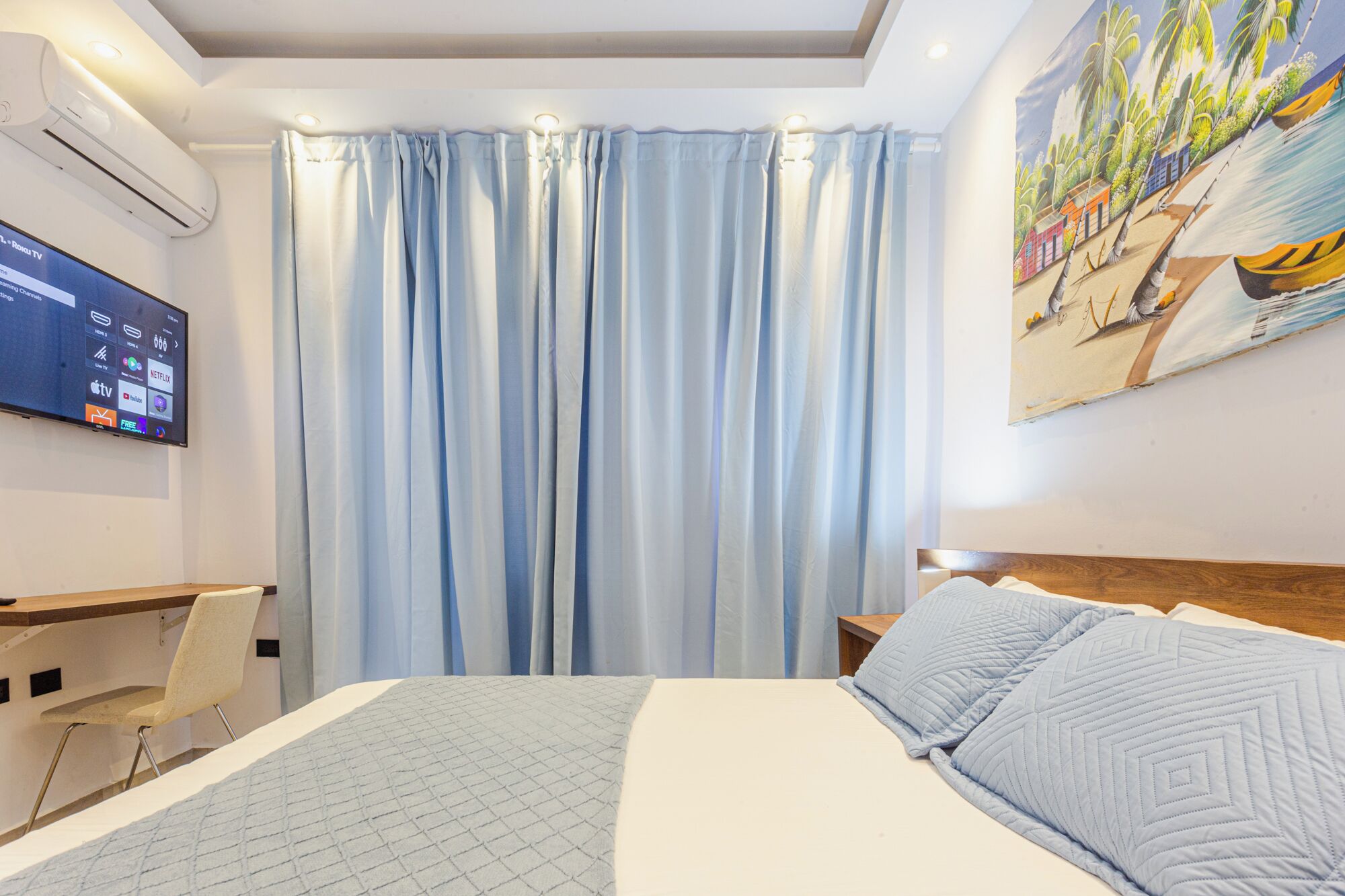 Una habitación de hotel con cama, escritorio, silla, televisor montado en la pared y un cuadro.