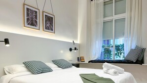 Una cama bien hecha con ropa blanca y almohadas a cuadros, una manta verde y un asiento junto a la ventana con vistas a la vegetación.
