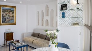 A living room with a sofa, a coffee table, a painting, and a decorative shelf with books and a bottle. - Estate4home- Casetta Arienzo (Positano)