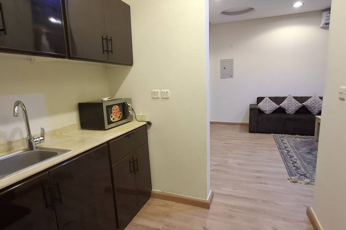 Una cocina compacta con gabinetes oscuros, un lavamanos y un microondas. Se aprecia una pequeña alfombra en la zona de estar contigua.