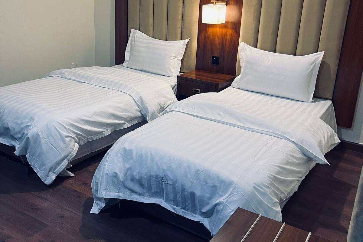 Habitación de hotel con dos camas individuales, cabecero de madera, mesitas de noche con lámparas y un banco de madera.