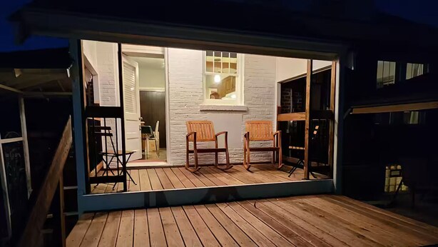 Una terraza de madera con dos mecedoras y vista a un cuarto con ventana y puerta.