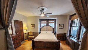 Un dormitorio con cama de madera, ventilador de techo, una silla, cómoda y ventana con cortinas.