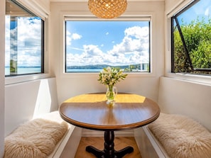 Una habitación de esquina con una mesa redonda de madera, un florero con flores y vistas al cielo y follaje a través de amplias ventanas.