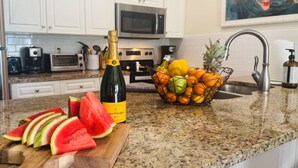 A kitchen with white cabinets, a microwave, a toaster, a bottle of champagne, a fruit basket, and a cutting board with watermelon slices. - Discounted GroundFloor. King Bed. Queen Bunks.Twin bunks. Wheelchair Accessible. (Orange Beach)
