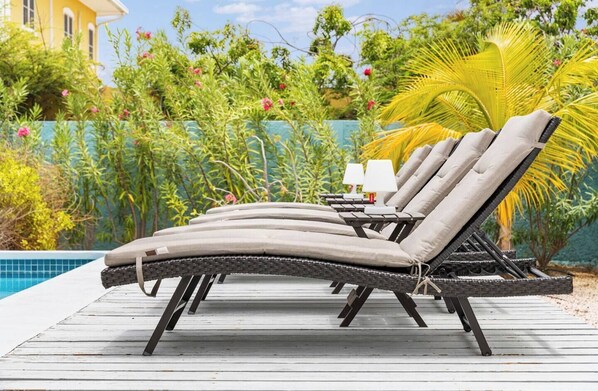 A row of outdoor lounge chairs with cushions and small lamps, positioned on a wooden deck by a pool, surrounded by lush greenery and a palm tree. - North end Villa with Magna Pool, Sea View, WiFi, AC, dive locker and rinse tanks (Kralendijk)
