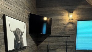 A room with wooden walls, a mounted TV, a framed artwork, a window with blinds, and a bed with a blanket. - Charming barn in Glasgow, modern amenities, private setting close to downtown (Glasgow)